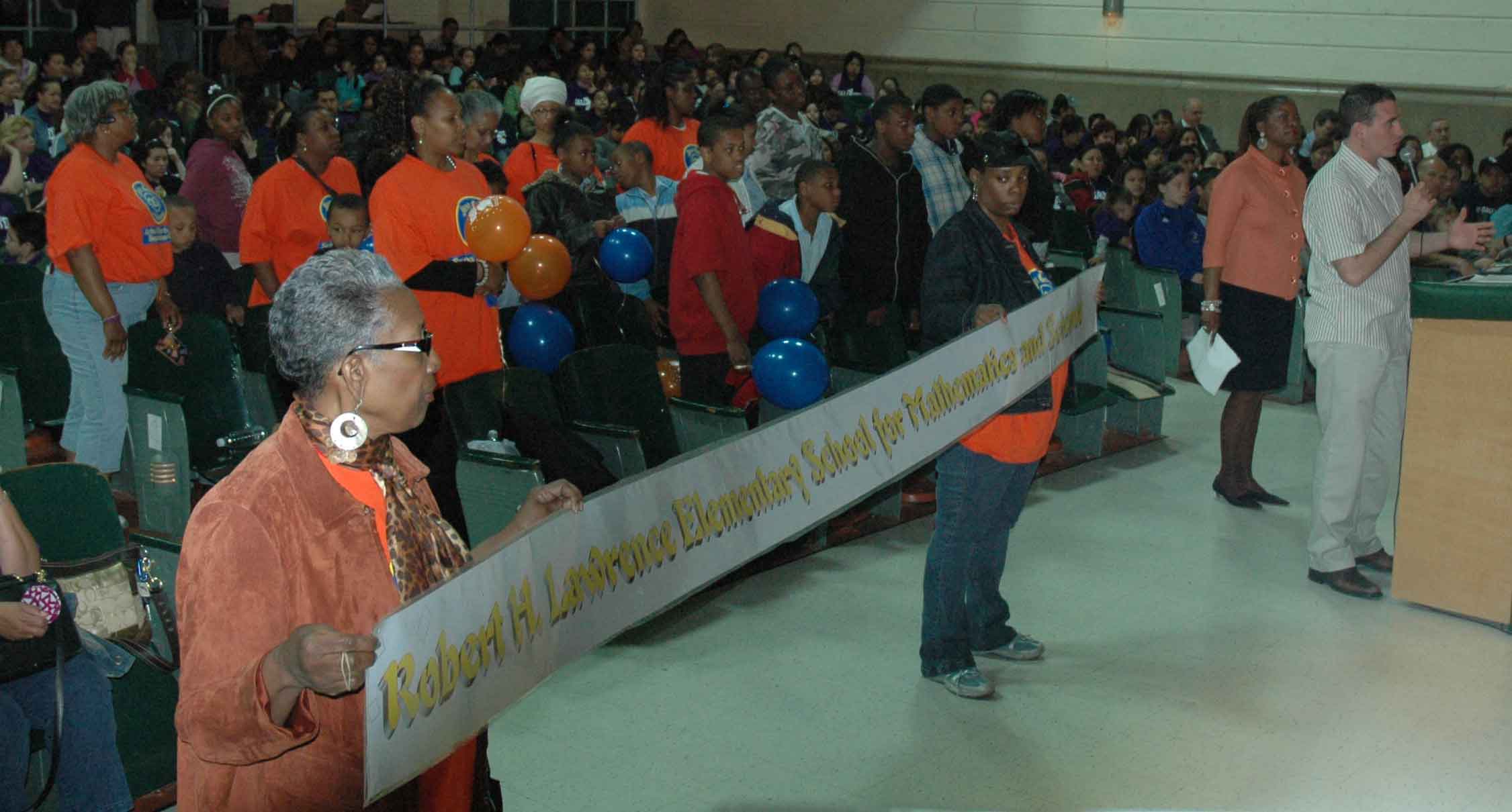 While their engineer Paul Casasanto delivers a detailed outline of the needs of their school (above right, standing with principal McClinton), parents and staff from Robert H. Lawrence Elementary stand during the May 22 hearing at Morgan Park High School. One of the newest trends at Chicago schools is that the school community designs a tee shirt when the school has to present its problems to the Board of Education. On May 22, Gallistel appeared in purple, while Lawrence was in red. Substance photo by George N. Schmidt.