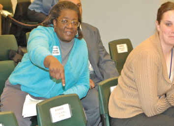 Packing the seats with administrators and “guests” keeps the public out of Board of Education meetings. Despite the fact that more than 250 people wanted to attend the March 28, 2007, meeting of the Chicago Board of Education, most were barred from the Board’s 5th floor meeting room and forced to spend the morning in “holding rooms” on the 19th floor. How? The Board reserves most of the seats in the room (especially those behind the speakers’ microphone where people will be seen on TV) for highly paid administrators and “guests.” Substance estimates that the administrators who spend the day every Board meeting taking up seats in the Board chambers have a total annual salary cost of more than $8 million. For more than ten years, it’s been possible to tell whether the Board is worried about the public participation session by noting the large number of young white women who fill seats directly in camera range. For reasons that have never been explained, the Board invites “social worker interns” to fill seats when it runs out of expensive administrators and political patronage appointees. Above: PURE’s Wanda Hopkins points to one of the seats reserved for the “social worker interns” before the beginning of the March 28 Board meeting. At the time Hopkins noted this, more than 150 people had been shuttled to the 19th floor “holding room” to keep them from the Board chambers. The social worker intern (right) did not identify herself. (Substance photo by George N. Schmidt).